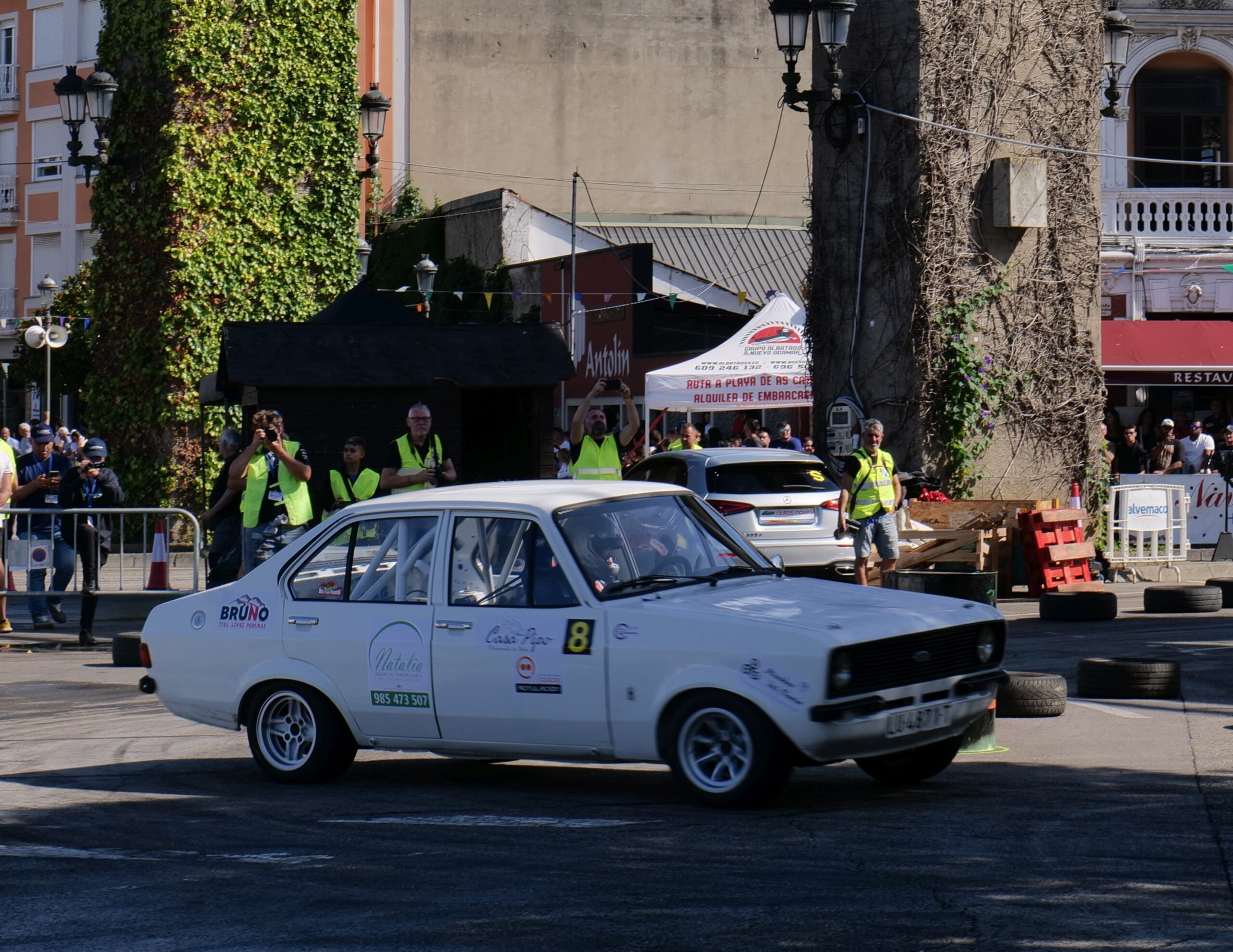 White rally car Ford turning on a city street during a motorsport event, with spectators and marshals nearby