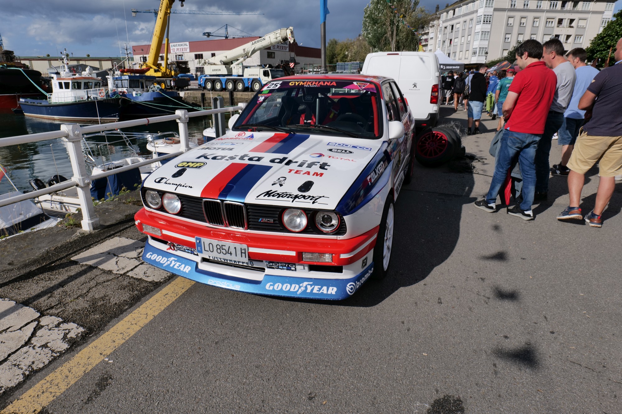 Rally car with sponsor decals parked by a harbor, with spectators nearby